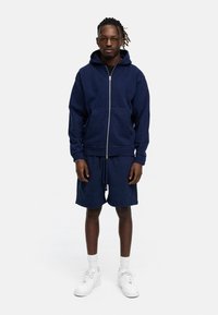 Navy blue zip-up hoodie with front pockets, paired with matching shorts. White sneakers and ribbed ankle socks complete the outfit.