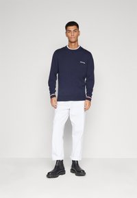 Maglione blu navy con polsini a costine, caratterizzato da un piccolo logo sul lato sinistro del petto; abbinato a pantaloni bianchi e stivali neri alla caviglia.