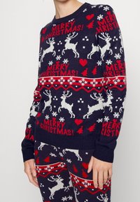 Mörkblå stickad jultröja med vita renar och röda detaljer, med snöflingor och texten "GOD JUL!" utspridd över hela.