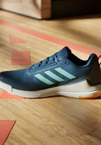 Scarpa da ginnastica blu scuro con strisce verde chiaro e suola bianca appoggiata su un pavimento di legno con righe rosse del campo.