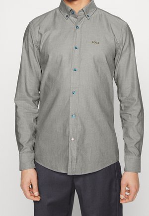 Homme portant une chemise grise à manches longues avec des boutons bleus et le logo "BOSS" sur la poitrine, associée à un pantalon sombre à rayures.