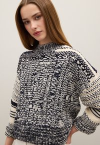 Grober Strickpullover in Marineblau und Creme. Verfügt über einen hohen Kragen, aufwendige Muster und überschnittene Schultern. Hergestellt aus strukturiertem Garn.