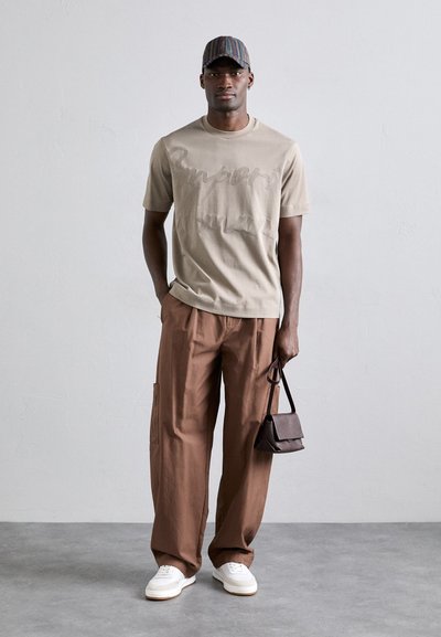 Beige bomull T-skjorte med subtil tekstdesign, brune vidde cargo-bukser, hvite joggesko og en stripete caps. Liten brun skulderveske.