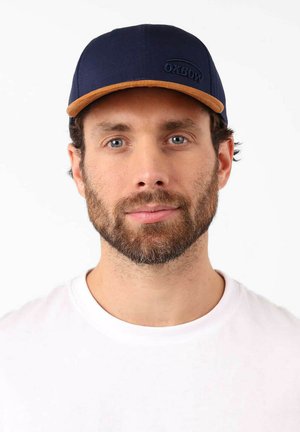 Casquette bleu marine avec une visière en cuir beige et le logo Oxbow. Le tissu est texturé, et le design présente une forme structurée avec un dos ajustable.