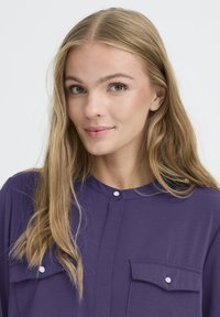 Vijolična srajca z gumbom, zaobljenega ovratnika, dvema sprednjima žepoma in mehkobo ter gladko teksturo. Ima bele gumbe spredaj.