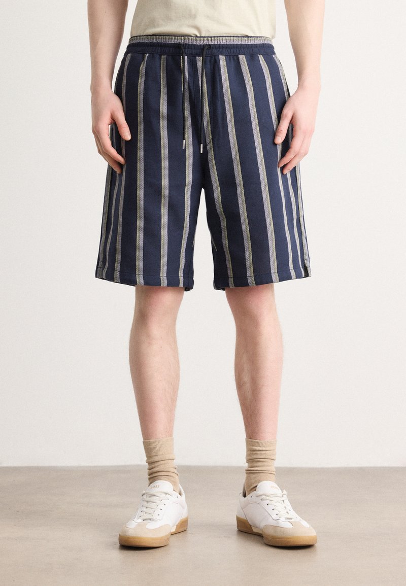 Shorts rayés navy en tissu léger, dotés d'une taille élastique à cordon de serrage et de poches latérales, complétés par des baskets blanches et des chaussettes beiges.