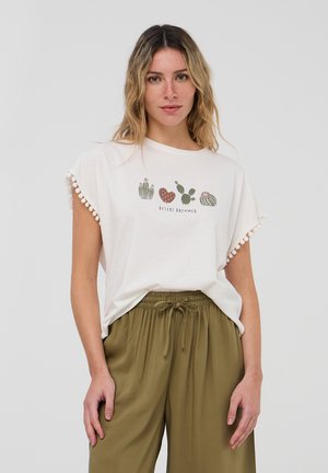 T-shirt bianco a maniche corte con grafica di cactus e testo "DESERT DREAMER", caratterizzato da rifinitura con pompon sulle maniche, abbinato a pantaloni verde oliva.