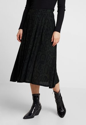 Femme portant une jupe midi à motif vert foncé, un haut noir à manches longues et des bottines noires brillantes avec des bouts pointus et des talons compensés.