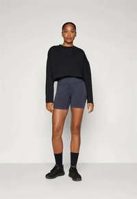 Fekete, rövidített pulóver hosszú ujjakkal, sötét szürke, testhez simuló rövidnadrággal párosítva. A modellen fekete sportcipő és zoknik vannak. Semleges háttér.