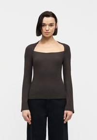 Γυναίκα με κοντά σκούρα μαλλιά, ντυμένη με εφαρμοστό μακρυμάνικο σκούρο καφέ τοπ με τετράγωνη λαιμόκοψη και μαύρο παντελόνι, που κοιτά μπροστά πάνω σε απλό φόντο.