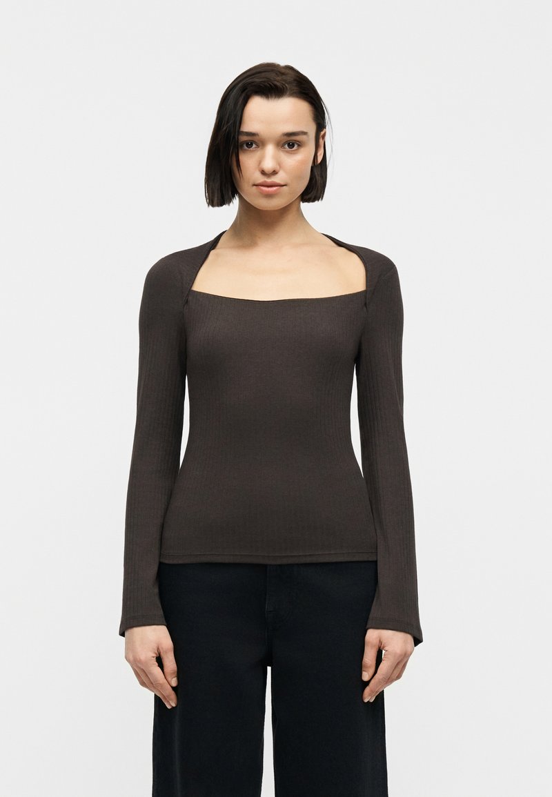 Γυναίκα με κοντά σκούρα μαλλιά, ντυμένη με εφαρμοστό μακρυμάνικο σκούρο καφέ τοπ με τετράγωνη λαιμόκοψη και μαύρο παντελόνι, που κοιτά μπροστά πάνω σε απλό φόντο.