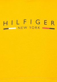 Geltona medvilninė džemperis su spausdintu "HILFIGER NEW YORK" logotipu tamsiai mėlynos, baltos ir raudonos spalvų, su apvalia iškirpte ir įtempimu apatinėje dalyje.
