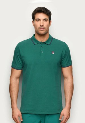 Hombre vestido con una camiseta polo verde de manga corta con un pequeño logo en el pecho izquierdo, de pie frente a un fondo claro y uniforme.