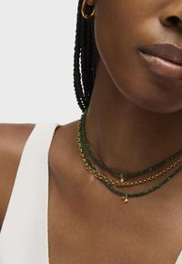 Collane a più piani con perle verdi, catena dorata e piccoli accenti in oro e perla. Pelle liscia con una maglietta bianca sotto.