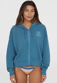 Sweatshirt à capuche bleu avec une fermeture éclair sur le devant et un design de logo graphique. Il présente une coupe décontractée et un tissu doux et texturé.