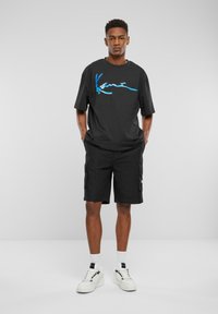 Sort oversized t-shirt med blåt grafisk design, kombineret med sorte cargo shorts og hvide sneakers. Modellen står med hænderne i lommerne.