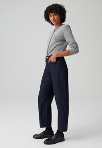 Cardigan grigio lavorato a maglia, jeans a gamba larga blu scuro e mocassini neri. I jeans presentano una vita alta, tasche frontali e dettagli di cucitura visibili.