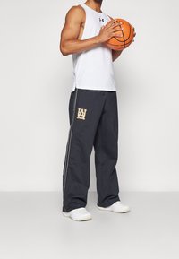 Grå träningstights med vita sidosträck och en logotyp, åtföljt av en vit ärmlös topp, hållande en orange basketboll.