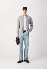 Cardigan a coste grigio chiaro, camicia bianca, jeans larghi blu, cintura nera, scarpe nere, mentre tiene una borsa compatta nera, in piedi su uno sfondo bianco.