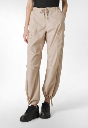 Beige cargo bukser med elastisk talje, sidelommer og strammede ankler, parret med sorte sneakers. Fremstillet af letvægtsmateriale.