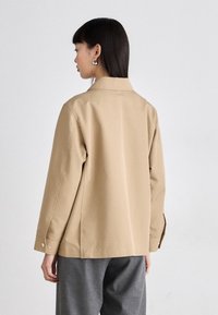 Light beige jacket with a collar, long sleeves, and side slits. Smooth fabric with a relaxed fit and minimalistic design. Silver button details.
