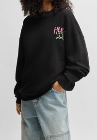Felpa nera con vestibilità ampia, caratterizzata da un logo grafico rosa e verde sul petto. Realizzata in materiale morbido, con polsini e orlo a costine.