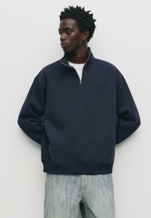 Felpa blu navy con zip, colletto alto e polsini a costine, abbinata a jeans in denim chiaro con lavaggio sbiadito. Design semplice e casual.