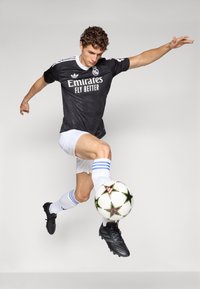 Male soccer player in black jersey and white shorts kicks a soccer ball mid-air against a plain background.