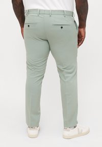 Άνδρας που φοράει ανοιχτοπράσινο παντελόνι στενής εφαρμογής και λευκά παπούτσια Veja, απεικονίζεται από πίσω μπροστά σε μια απλή φόντο.