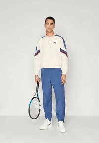 White zip-up track jacket with blue and red accents, paired with blue track pants and white sneakers, holding a tennis racquet.