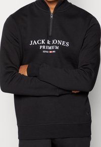 Czarna rozpinana bluza z wysokim kołnierzem, z białym napisem "JACK & JONES PREMIUM" oraz logo na dolnym prawym boku. Gładka faktura.