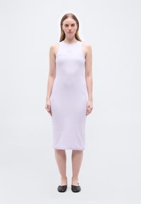 Γυναίκα που στέκεται και κοιτάζει μπροστά φορώντας ένα αμάνικο midi φόρεμα σε ανοιχτό μοβ χρώμα και μαύρα επίπεδα παπούτσια σε απλό ανοιχτό φόντο.