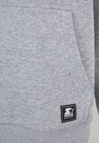 Sudadera gris con un tejido texturizado, que presenta un bolsillo frontal y una etiqueta negra con un logo de estrella blanca en el lado inferior.