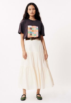 T-shirt graphique noir avec un texte coloré, associé à une jupe longue blanche à volants. Porté avec une ceinture marron et des chaussures slip-on vertes.