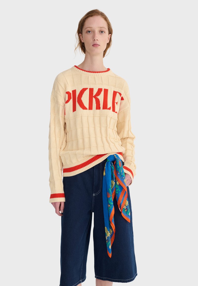 Jeune femme portant un pull crème avec le texte rouge « PICKLES », un pantalon ample bleu foncé, et une écharpe fleurie bleue nouée à la taille.