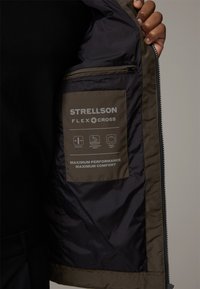 Olivengrøn jakke med lynlås, udstyret med et glat inderskær og et mærke mærket "STRELLSON FLEX CROSS" med ydeevnedetaljer.