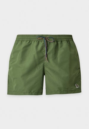 Homme torse nu aux cheveux foncés portant un short de bain vert à cordon, debout pieds nus sur un fond clair uni.