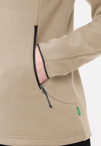 Beige Fleecejacke mit seitlichem Reißverschluss, schwarzem Besatz und Kordelzug. Verfügt über einen strukturierten Stoff mit glatter Oberfläche und flachen Nähten.