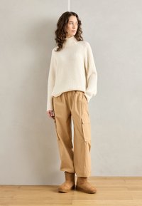 Crème geribbelde turtleneck trui, baggy beige cargobroek met zijzakken en tan enkellaarzen, staande op licht houten vloeren.