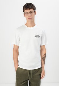 T-shirt bianco in cotone con scollatura rotonda, maniche corte e logo stampato nero sul petto sinistro. Il modello indossa pantaloni in velluto a coste verde oliva.