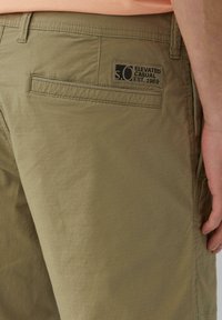 Pantaloni corti verde oliva realizzati in tessuto leggero, con una tasca posteriore con etichetta nera, cuciture decorative e una vestibilità comoda.