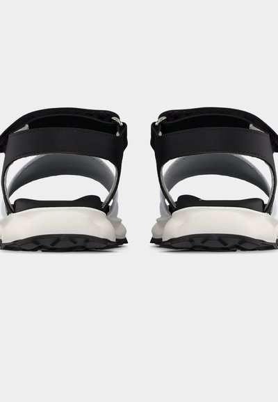 Ζευγάρι μαύρες και λευκές σαγιονάρες με ρυθμιζόμενους μαύρους ιμάντες και χοντρές λευκές σόλες, που φαίνονται από πίσω σε απλό φόντο.