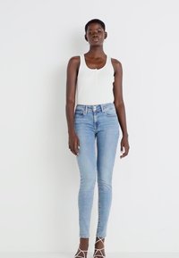 Top corto de tirantes blanco acanalado y jeans ajustados de color azul claro. Los jeans tienen una cintura alta y un ligero desgaste. Modelo lleva tacones nude con tiras.