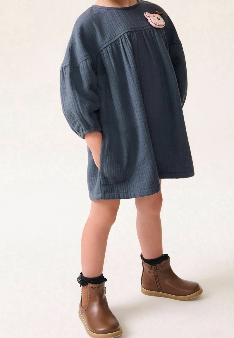 Enfant portant une robe ample gris foncé avec un patch animal rose et des bottines marron avec des chaussettes noires en dentelle, debout les mains dans les poches.