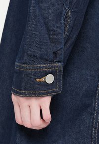 Manche de veste en denim bleu foncé avec des coutures jaunes contrastantes, un bouton-pression en argent et une légère texture. Main visible au bord.