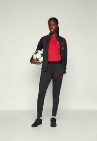 Mustera mustroniga jakk punaste aktsentidega, punane särg ja mustad püksid. Hoides musta ja valget jalgpalli, millel on Adidas logo.