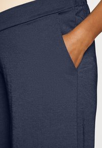 Calças de tecido azul marinho com uma textura suave, apresentando um bolso lateral e uma cintura elástica para conforto. Detalhe em close da área do bolso.