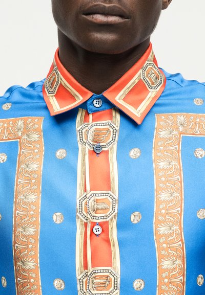 Prim-plan cu o persoană purtând o cămașă albastră și portocalie cu modele geometrice și botanice, având guler cu nasturi și pată frontală.