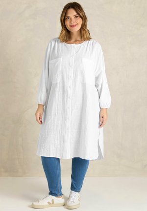 Kvinde iført en lang hvid kjole med knapper og lommer, blå jeans og hvide sneakers, stående mod en beige baggrund.