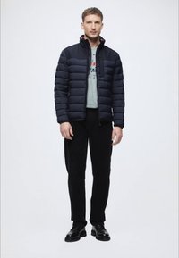 Sötétkék pufferekő kabát magas gallérral, cipzáros záródással és finom varrásokkal. Szürke ing fölött viselve, feketére szabott nadrággal és cipővel párosítva.